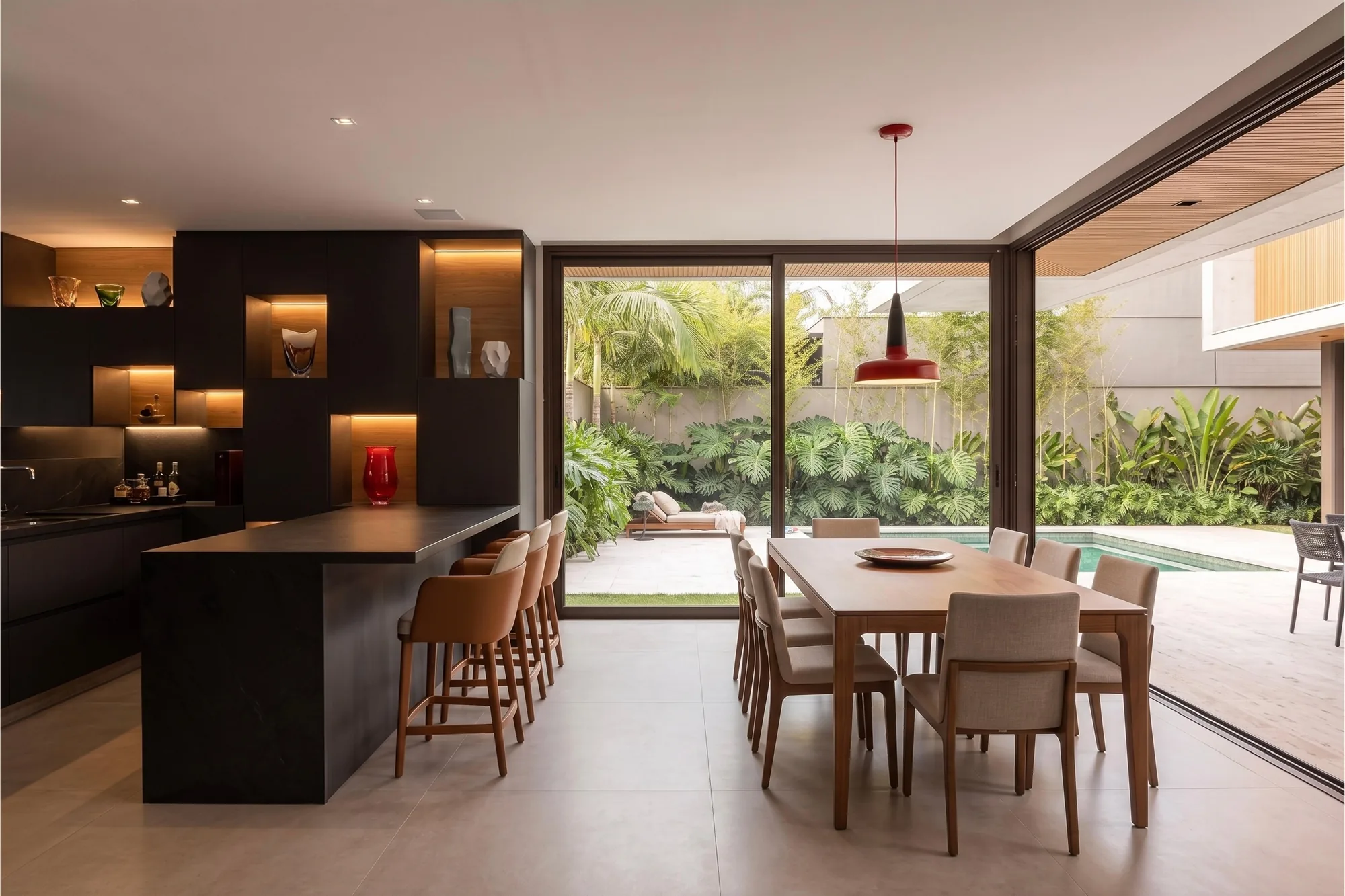 Cozinha integrada à sala de jantar com marcenaria preta, nichos iluminados e pendente vermelho sobre mesa de madeira, com piscina verde ao fundo